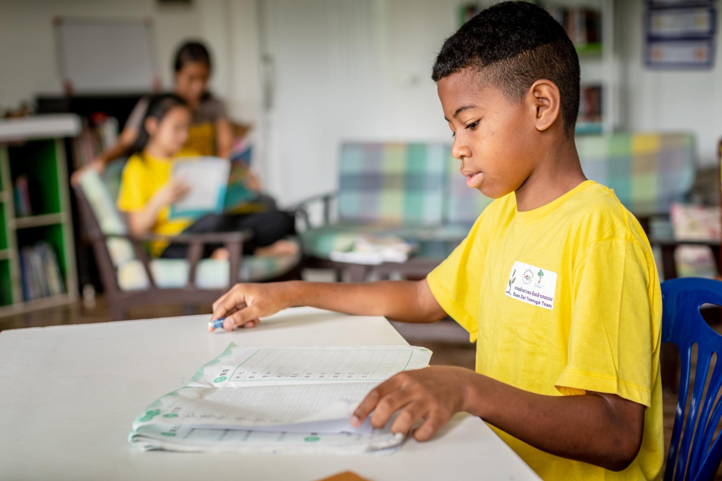 Ein Junge aus Thailand sitzt an einem Tisch und liest in einem Arbeitsheft (Quelle: KNH / Jakob Studnar)